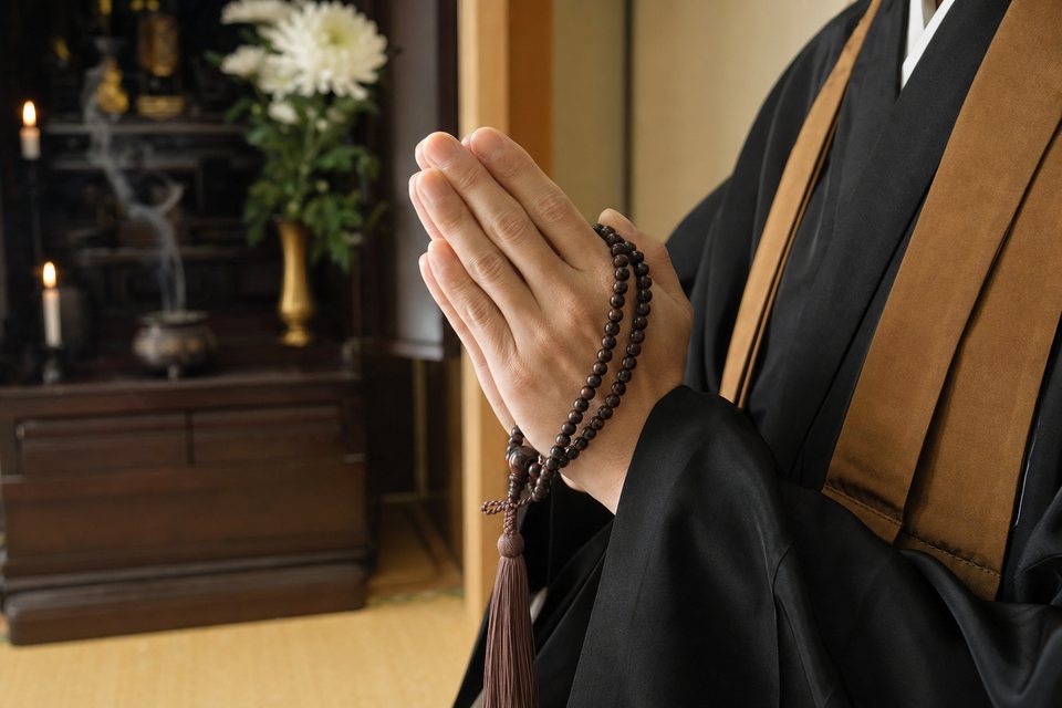 数珠を手に合掌する僧侶の手元と、仏壇(イメージ)